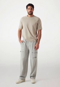 Beige T-shirt met korte raglanmouwen, gecombineerd met lichtgrijze cargobroek met twee ritssluitingen en bedrukte tekst. Witte sneakers.