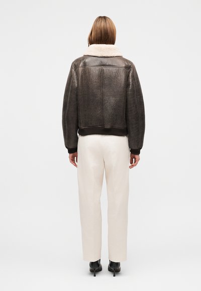 Καφέ δερμάτινο μπουφάν τύπου bomber με γιακά από shearling, ριμπ μανσέτες και κάτω μέρος. Φορεμένο με παντελόνι σε κρεμ απόχρωση και μαύρα παπούτσια με τακούνι.