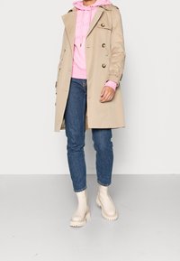 Gabardina beige con diseño de doble botonadura, hombreras, llevada sobre una sudadera con capucha rosa, combinada con vaqueros azules y botas color crema.