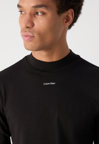 Must mustriga särk kõrge kaelusega, valmistatud pehmest kangast. Rinna peal on väike valge "Calvin Klein" logo. Sile tekstuur.