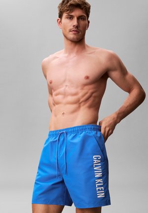 Man zonder shirt met strakke buikspieren, gekleed in blauwe Calvin Klein zwemshorts, tegen een effen grijze achtergrond, handen rustend nabij de zakken van de shorts.