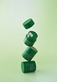 Trzy zielone cylindryczne słoiki do pielęgnacji skóry, o gładkiej fakturze i pasujących wieczkach, mają białe etykiety zawierające informacje o produkcie. Unoszą się na jasnoszarym tle.