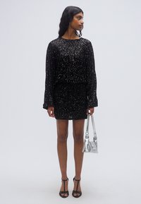 Μαύρο φόρεμα με παγιέτες, μακριά μανίκια, εφαρμοστή μέση και μήκος μίνι. Συνδυασμένο με μαύρα ψηλοτάκουνα με λουράκια και ασημένια τσάντα χειρός.