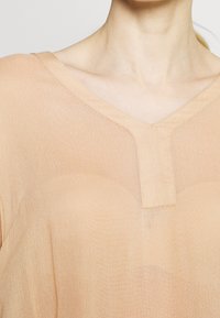 Blouse légère de couleur pêche avec un décolleté en V et des manches courtes, dotée d'une texture légèrement transparente et d'un détail sur le panneau central.