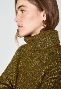 Maglione chunky di colore verde oliva con colletto alto a coste, caratterizzato da motivi testurizzati e un filato morbido e spesso. Orecchini a cerchio dorati in accentuazione.
