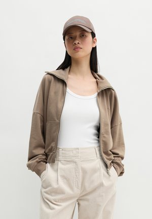 Jonge vrouw met lang donker haar, die een beige jas met rits, wit hemdje, beige ribfluwelen broek en een beige baseballpet draagt.
