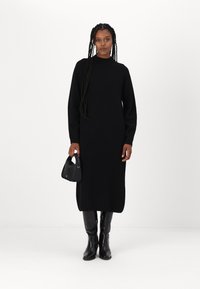 Abito nero in maglia con scollatura alta e maniche lunghe, caratterizzato da una vestibilità dritta e un orlo a lunghezza media. Accentuato da una piccola borsa nera.