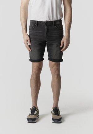 Mann trägt schwarze hochgekrempelte Jeansshorts, weißes T-Shirt und beige-schwarze Turnschuhe und steht vor einem schlichten grauen Hintergrund.