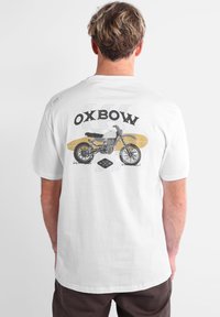 Persona che indossa una maglietta bianca con una grafica di una motocicletta e una tavola da surf, con la scritta "OXBOW" stampata sul retro.