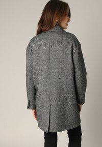 Grijze visgraatmantel met een gestructureerde silhoetten, opstaande kraag en zijsplitten; textuurstof met subtiele zilveren spikkels, lange mouwen.