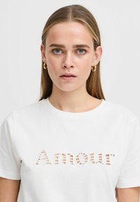 Bílý bavlněný tričko s výstřihem zřetelným nápisem "Amour" v vystřiženém designu se pruhovaným vzorem. Kulatý výstřih a krátké rukávy. Měkká textura.