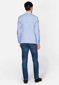 Jasnoniebieska marynarka w kratkę z gładkiego materiału, z jedną rozcięciem z tyłu, noszona na ciemnych dżinsach i brązowych butach. Widoczne detale guzików na mankietach.