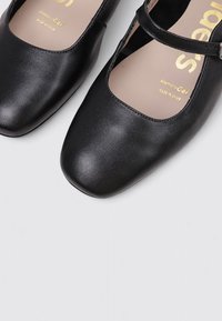 Zapatos Mary Jane de cuero negro con puntera cuadrada, plantillas de gel con memoria y una correa ajustable con hebilla sobre un fondo gris.
