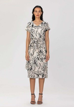 Femme portant une robe beige jusqu'aux genoux, à manches courtes, avec des motifs floraux abstraits noirs et des talons noirs à bride à la cheville, debout devant un fond uni.