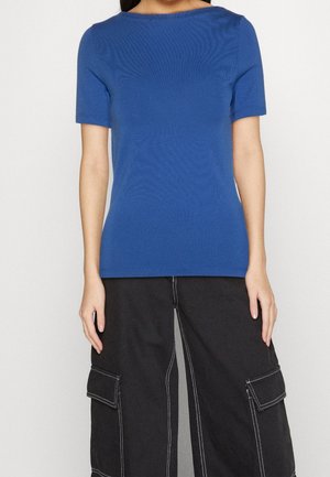 Woman wearing a fitted blue short-sleeve shirt and loose black cargo pants with white stitching against a plain background.