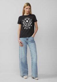 Zwarte katoenen t-shirt met een witte florale grafische print en tekst. Gecombineerd met lichtblauwe wijdvallende jeans en lichtgekleurde schoenen.