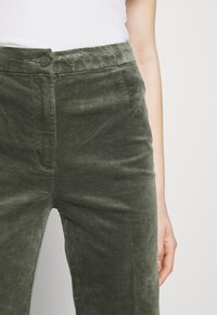 Personne portant un pantalon en velours côtelé vert olive taille haute et un haut blanc côtelé, la main posée à côté du pantalon.