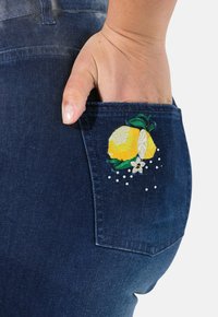 Tmavě modré džíny s rukou v kapse, zdobené živou výšivkou citronů a listů s dekorativními korálky.