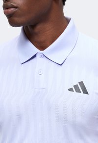 Gros plan d'une personne portant un polo blanc Adidas avec un motif texturé et un logo noir sur la poitrine.