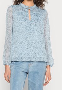 Blouse florale bleu clair avec de longues manches transparentes, un décolleté clé et un détail de lien. Tissu doux et texturé avec un motif délicat.