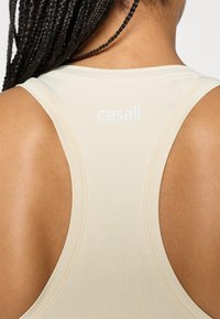 Κοντινό πλάνο ενός ατόμου με πλεγμένα μαλλιά που φοράει ένα μπεζ κοντομάνικο τοπ με λογότυπο "casall" στην πάνω πλάτη.