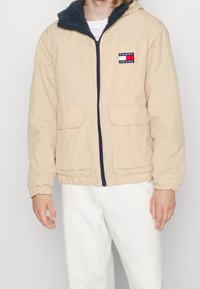 Beige puffer-jakke med en marineblå lynlås, fleece-foring i hætten og to forlommer. Har et logo-patch på venstre brystområde.