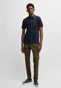 Marinblå polo skjorta med gult-grönt kantband, klassisk krage, tvåknapps knäppning, i kombination med olivgröna byxor och marinblå sneakers.