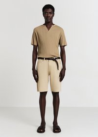 Camicetta beige a righe con maniche corte e scollatura a V, abbinata a pantaloni corti beige. Una cintura intrecciata scura e sandali neri completano l'outfit.