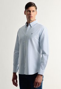 Camisa de manga larga a rayas azul claro con cuello abotonado y logo bordado en el pecho. Textura suave y ajuste entallado.