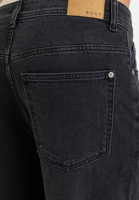 Jeans de mezclilla negra con un primer plano del bolsillo trasero, con una etiqueta de cuero estampada con "ROXY" y detalles de costura visibles.