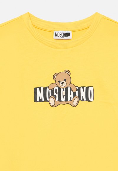 Rumena otroška majica z okroglim izrezom, na sprednji strani je medvedek, ki sedi nad krepko črno-belo napisano besedo "Moschino".