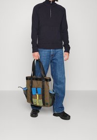 Canvasbärväska med gröna, blå och bruna sektioner, med svarta remmar, en framlapp och en gul accentdragkedja.