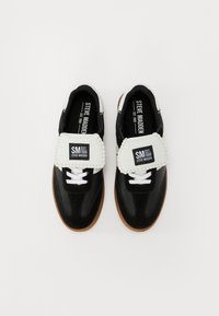 Schwarz-weiße Sneakers mit glänzender Oberfläche, ausgestattet mit einem gepolsterten Riemen, weißen Schnürsenkeln und einer braunen Gummisohle. Branding auf dem Riemen.