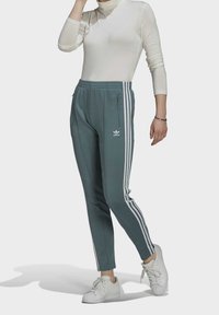 Pantaloni sportivi verdi con tre strisce bianche sui lati, dotati di vita con cordino e tasche con zip, abbinati a sneaker bianche.