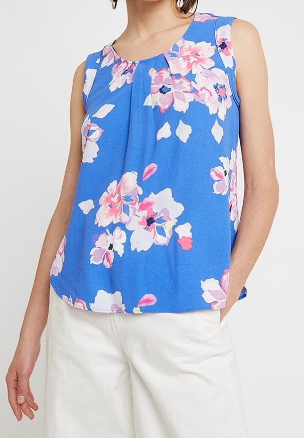 Haut bleu sans manches avec imprimé floral rose et blanc, porté avec un pantalon blanc, montré sur une personne avec la main dans la poche.