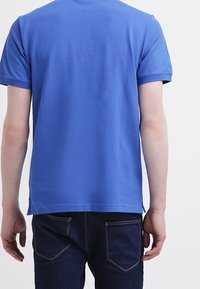 Camiseta de algodón azul con mangas cortas, cuello redondo y bajo recto. Llevada con jeans oscuros que tienen bolsillos traseros y costuras.