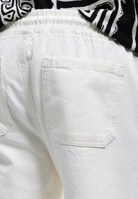 Pantalones de mezclilla blancos con cintura elástica. Presentan dos bolsillos traseros y una superficie texturizada. Parcialmente cubiertos por una camisa negra con estampado.