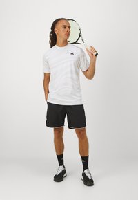 Valge lühikeste varrukatega spordisärk lainelise tekstuuriga, must logo, koos mustade lühikesete pükstega valgete aktsentidega. Hoides tennisereketit.