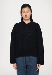 Maglione lavorato a maglia blu navy con scollo a V, motivo zigzag testurizzato, maniche ampie e orlo a coste, abbinato a jeans blu dalla vestibilità morbida.