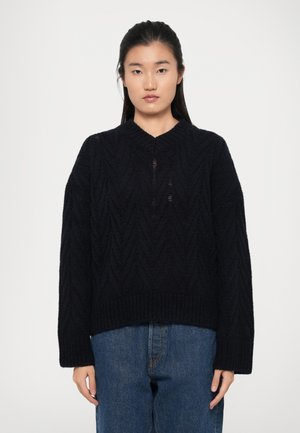 Mørkeblå strikkegenser med V-hals, teksturert zigzag-mønster, vide ermer og ribbestrikket kant, kombinert med avslappede blå jeans.