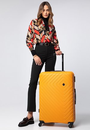 Yellow hardshell suitcase with a diamond pattern, four wheels, and a telescopic handle; paired with a multicolored patterned blouse and black pants.