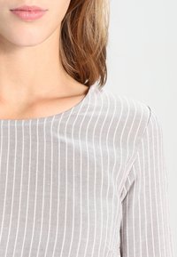Grå stribet top lavet af blødt stof med bådudskæring, der har vandrette striber og en glat tekstur. Nærbillede fremhæver design detaljer.
