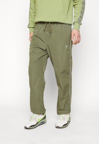 Olivově zelené větruvzdorné kalhoty s volným střihem, pasem na šňůrku, bočními pruhy a reflexním logem Nike na levé nohavici.