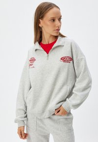 Sudadera gris clara con media cremallera, con texto rojo bordado, puños acanalados y bolsillos laterales. Combinada con una parte superior roja debajo.