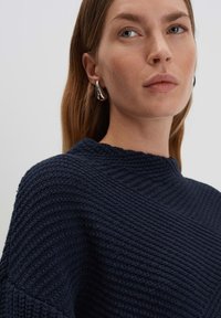 Maglione lavorato a maglia navy con colletto alto e motivo a coste diagonali. Il modello indossa orecchini a cerchio argentati. Design morbido e dal fit rilassato.