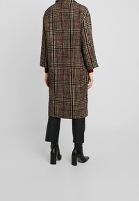 Cappotto a quadri in misto lana beige e nero, con dettagli rossi, maniche ampie e una vestibilità lunga e morbida, abbinato a pantaloni neri e tacchi.