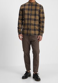 Camisa de cuadros marrón con cuello de banda, combinada con pantalones slim-fit marrón oscuro y zapatos negros con cordones.