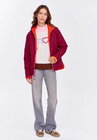 Kobieta w różowo-czerwonej kurtce, białym swetrze z niebieskim i czerwonym wzorem serc, szarych dżinsach i trampkach w panterkę, stojąca na białym tle.