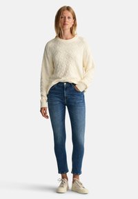 Gräddfärgad stickad tröja med diamantmönster, kombinerad med mörkblå slim fit-jeans. Modellen bär ljusa sneakers och visar en avslappnad outfit.