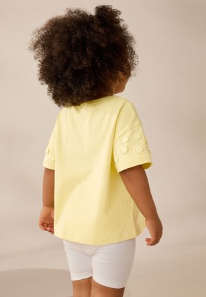 Bambino con capelli ricci che indossa una maglietta gialla a maniche corte con motivi floreali sulle maniche e pantaloncini bianchi aderenti, di spalle.